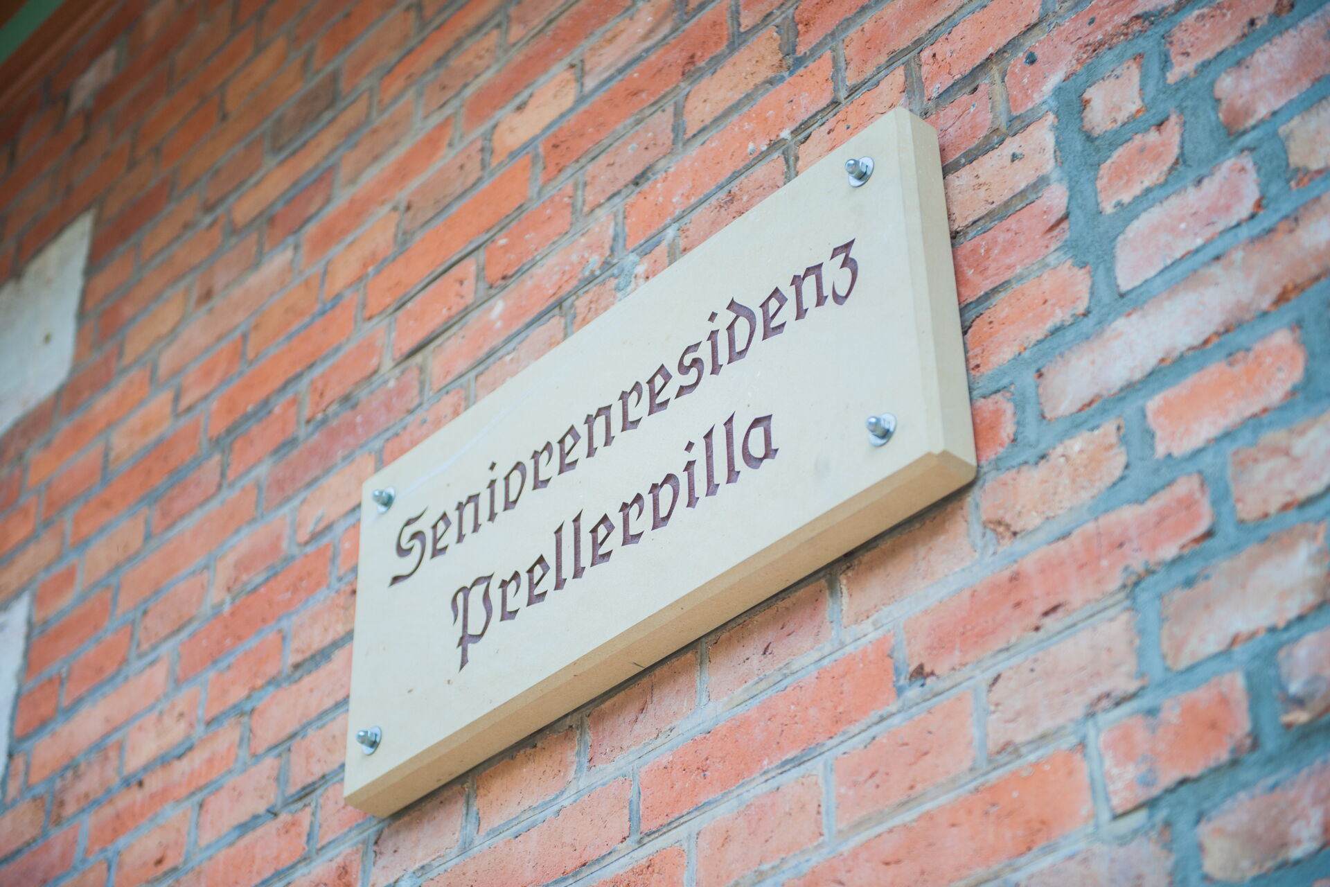 Ein helles Holzschild mit der Aufschrift "Seniorenresidenz Prellervilla" in alter deutscher Schrift, befestigt an einer roten Ziegelwand.