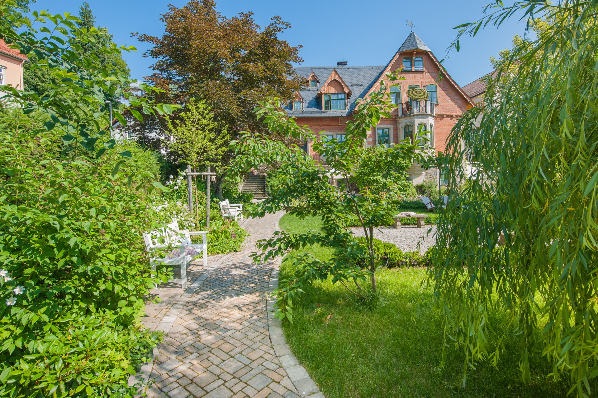 Ein idyllischer Garten mit einem geschwungenen gepflasterten Weg, der zu einer historischen Villa mit roter Backsteinfassade führt. Im Garten stehen weiße Bänke, üppige grüne Pflanzen und Bäume.