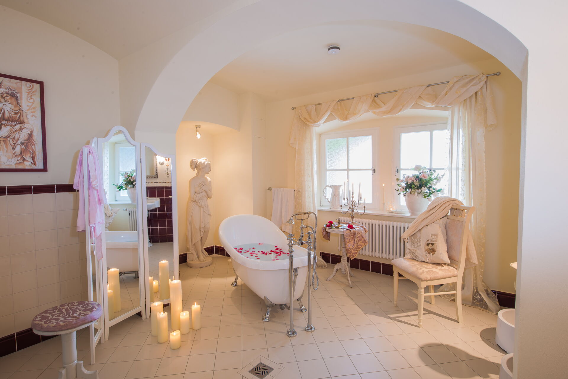 Ein luxuriöses, helles Badezimmer mit einer freistehenden Badewanne, gefüllt mit Wasser und roten Rosenblättern. Mehrere Kerzen in verschiedenen Größen brennen auf dem Boden.