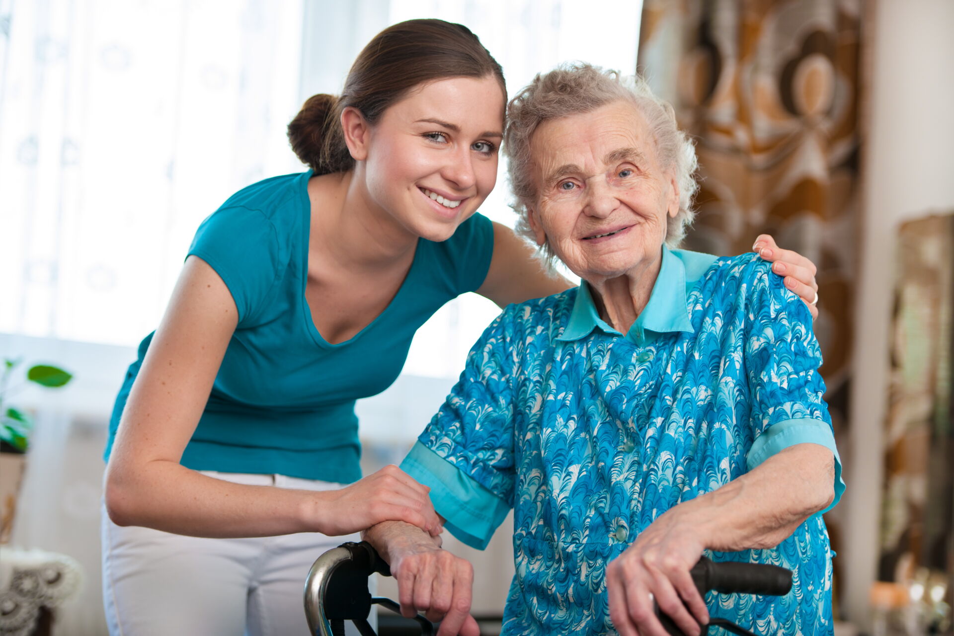 Eine junge, lächelnde Pflegekraft steht hinter einer älteren, ebenfalls lächelnden Seniorin, die sich auf einen Rollator stützt. Beide blicken freundlich in die Kamera.