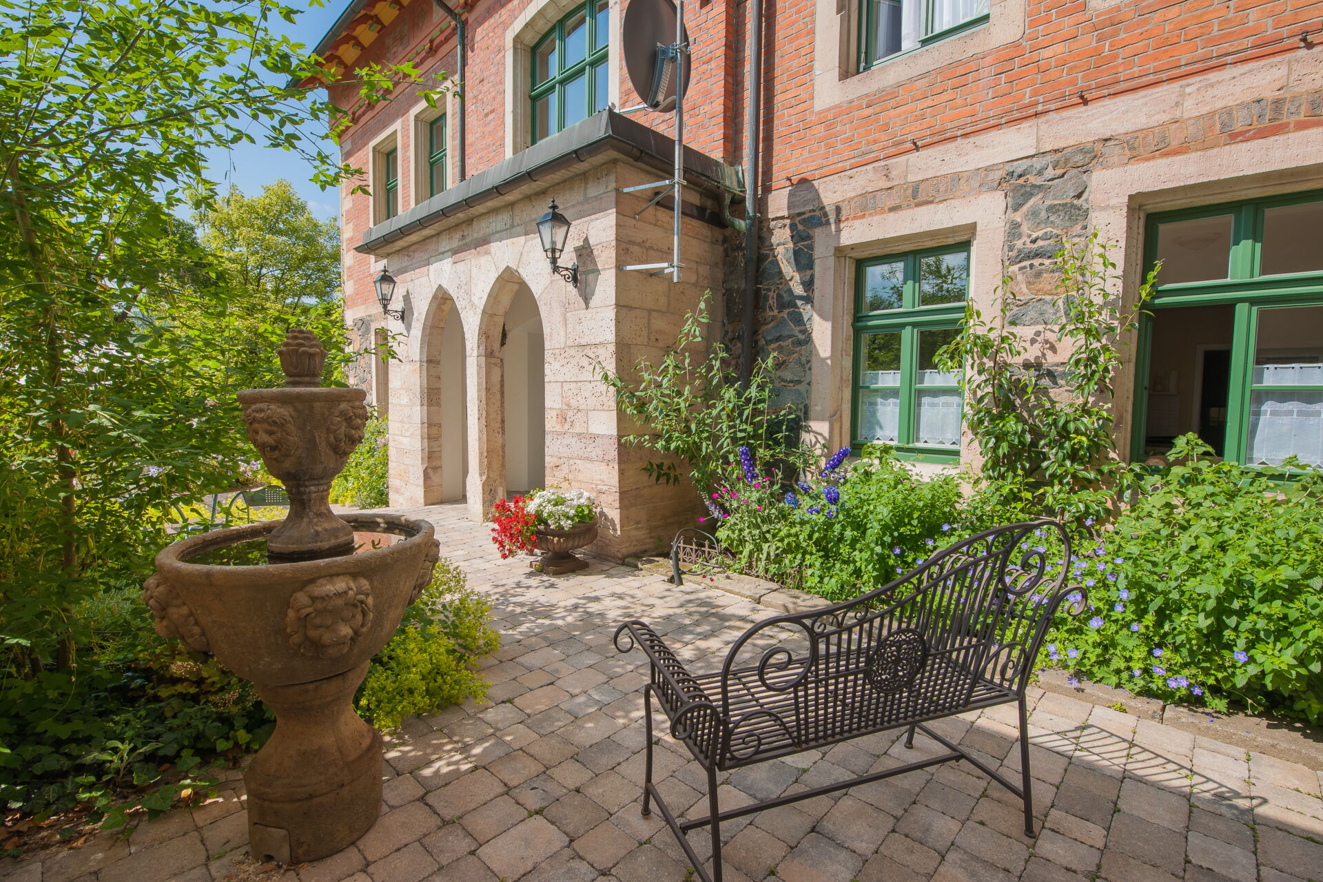 Ein gepflasterter Innenhof vor einer historischen roten Backsteinvilla mit grünen Fenstern. Im Vordergrund steht ein verzierter steinerner Brunnen und eine dunkle Metallbank.