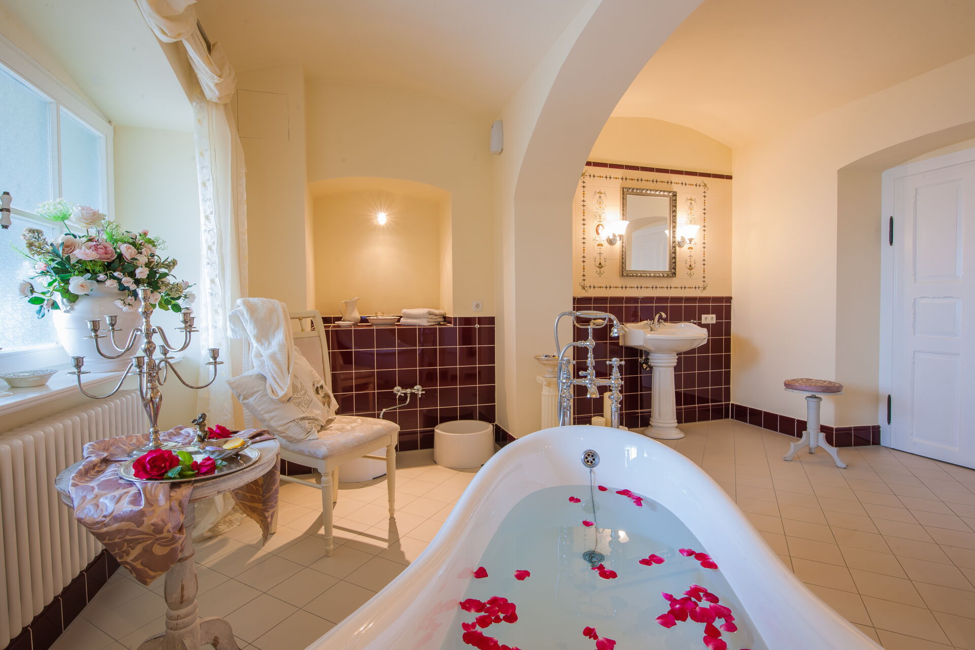 Ein stilvolles, geräumiges Badezimmer in einer Villa mit einer freistehenden weißen Badewanne, gefüllt mit Wasser und roten Rosenblättern.