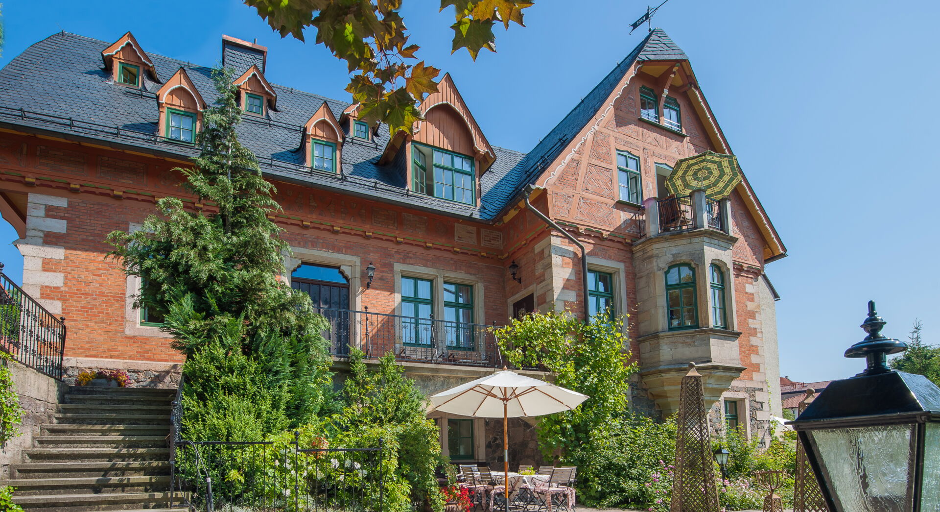 Eine prächtige, alte Villa mit roter Backsteinfassade, Fachwerkelementen und Schieferdach. Im Vordergrund befindet sich ein gepflasterter Hof mit einem Sonnenschirm und Gartenmöbeln. Rechts im Bild steht eine historische Laterne.